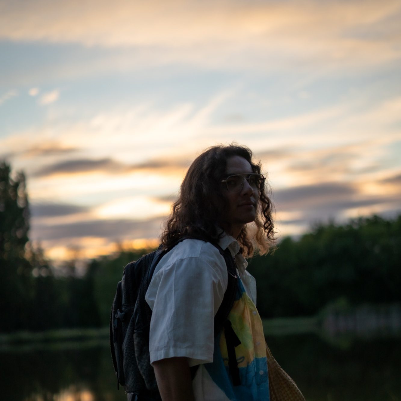 Un jeune homme avec des cheveux longs se tient près d'un lac au coucher du soleil.